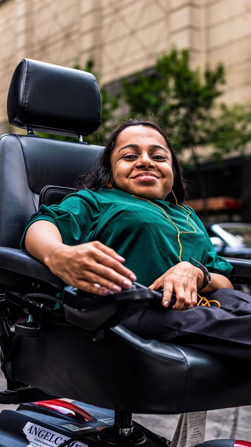 A young professional women with a big smile who is a power wheelchair user is crossing the street in a busy city.